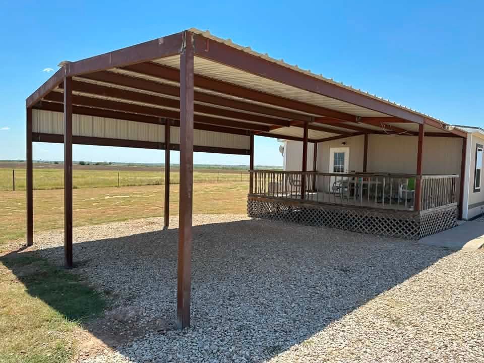 Rusty metal carport beside a white house, set on gravel in a rural field.