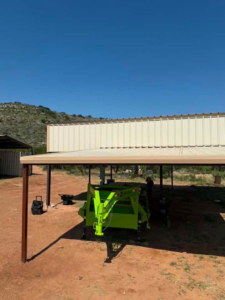 Green aircraft under a carport beside a tan building, on a red dirt lot under a clear blue sky
