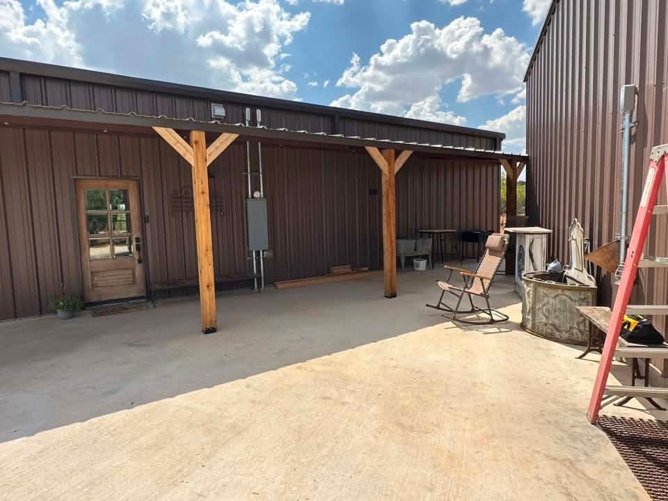 Covered patio beside a brown metal building with wooden posts, concrete floor, and outdoor chairs under a blue sky