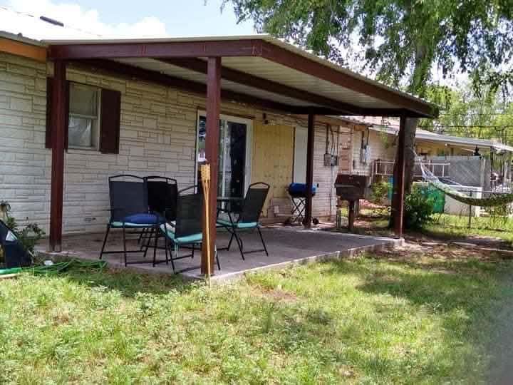 Covered patio with chairs and a grill beside a brick house, overlooking a grassy yard.