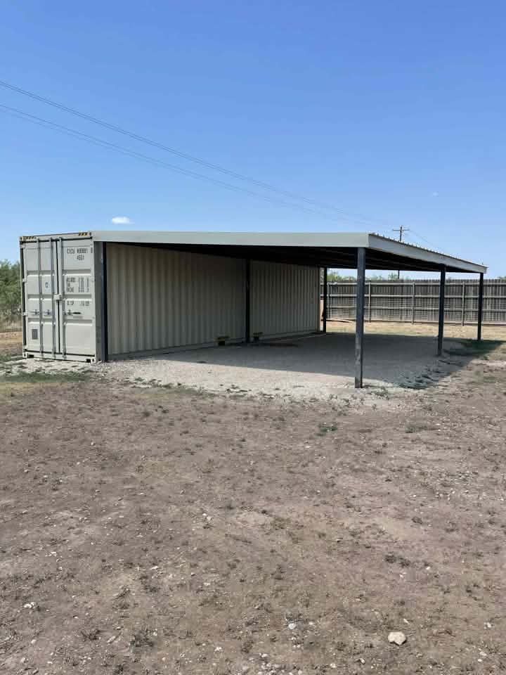 Open-sided metal carport with a shipping container wall, in a dusty outdoor lot under a clear sky