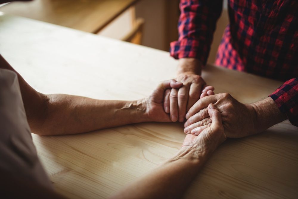 Hands clasped in support on a wooden table, with a person in a red plaid shirt nearby.