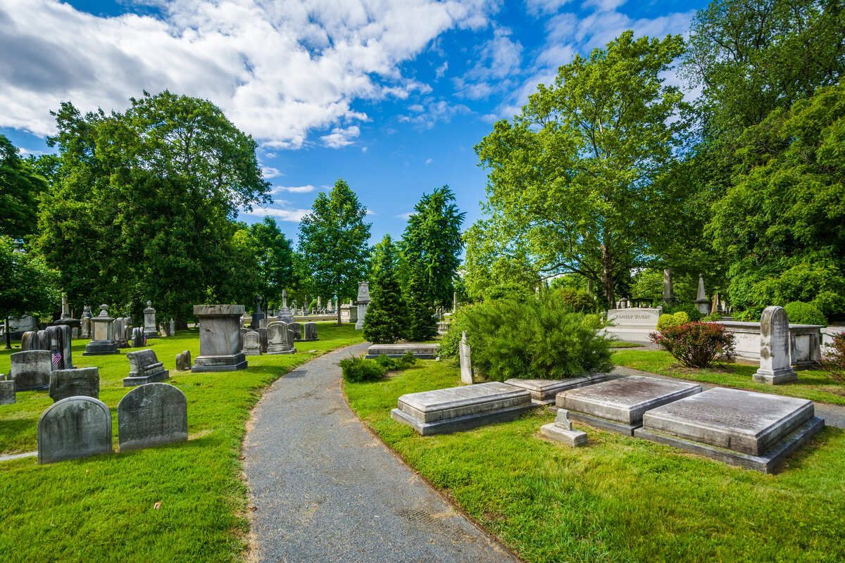 Curved path through a green cemetery with gravestones, trees, and a bright blue sky with clouds