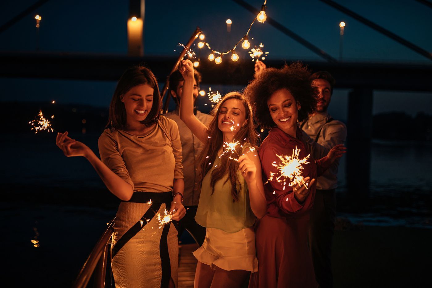 Three people smiling and holding sparklers under string lights at a nighttime party
