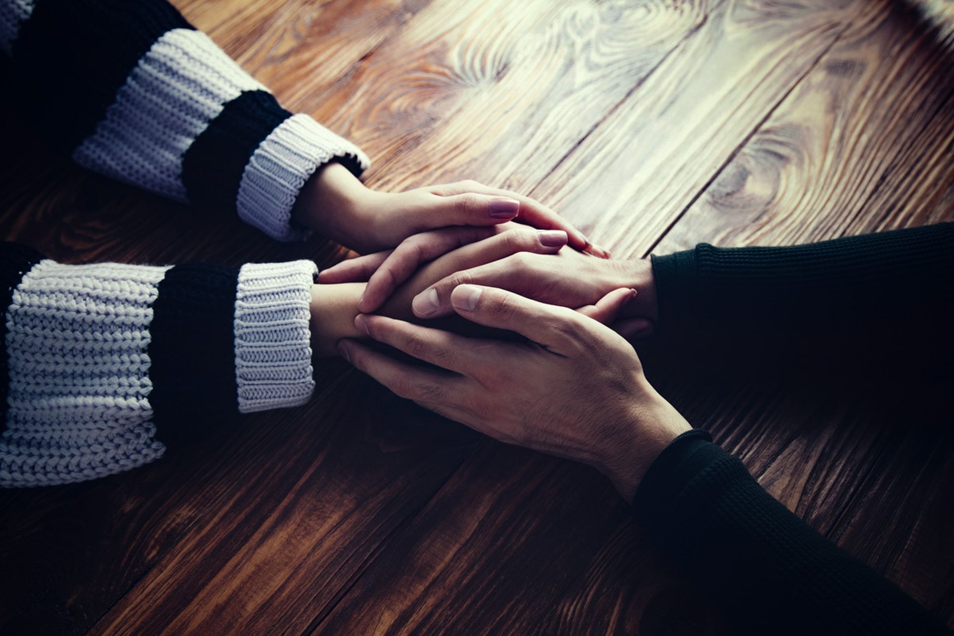 Two people holding hands across a wooden table, one in a black-and-white sweater and one in a black sleeve.