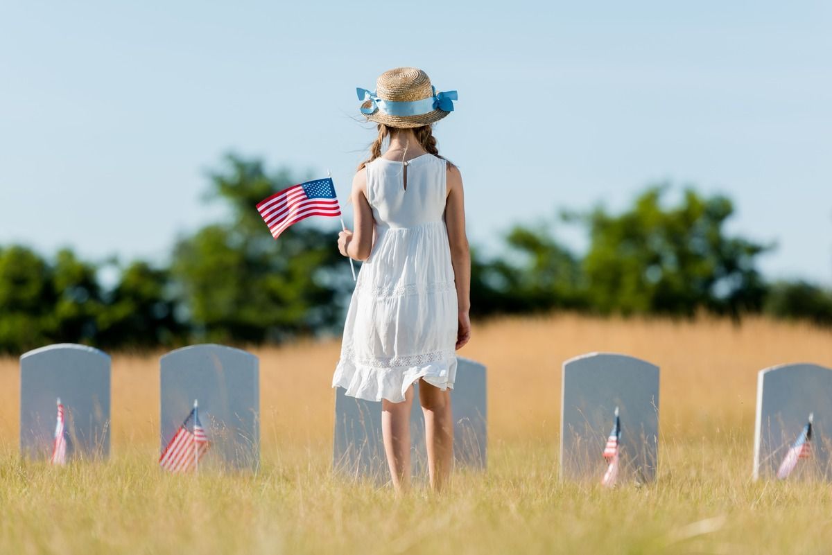 Child in a white dress and straw hat stands by military gravestones, holding a small American flag.