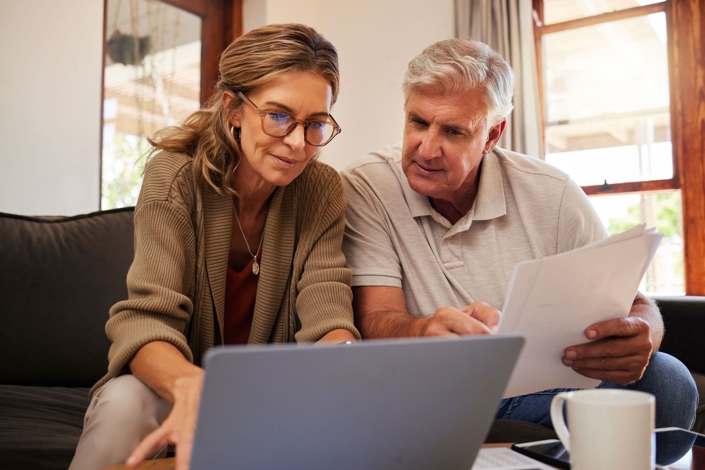 Two people review papers and a laptop at a table in a sunlit living room