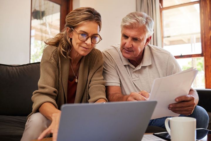 Two people review papers and a laptop at a table in a sunlit living room