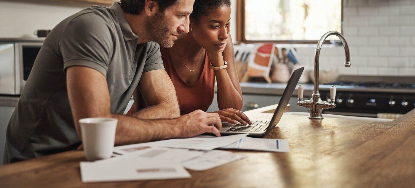 Two people leaning over a laptop at a kitchen table, reviewing paperwork in a sunlit home kitchen.