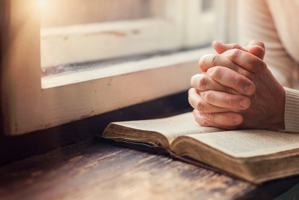 Praying hands resting on an open book by a sunlit window