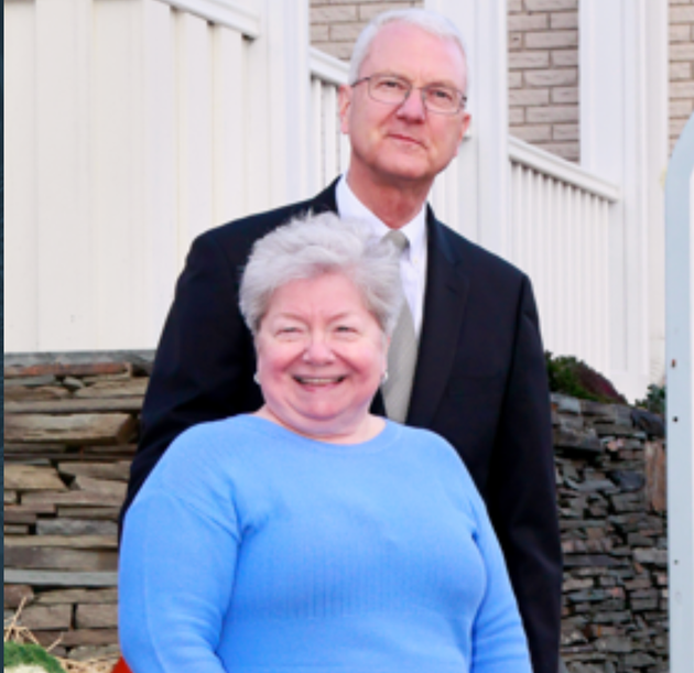 Smiling couple standing on steps outside a house, with a white railing and stone wall behind them.