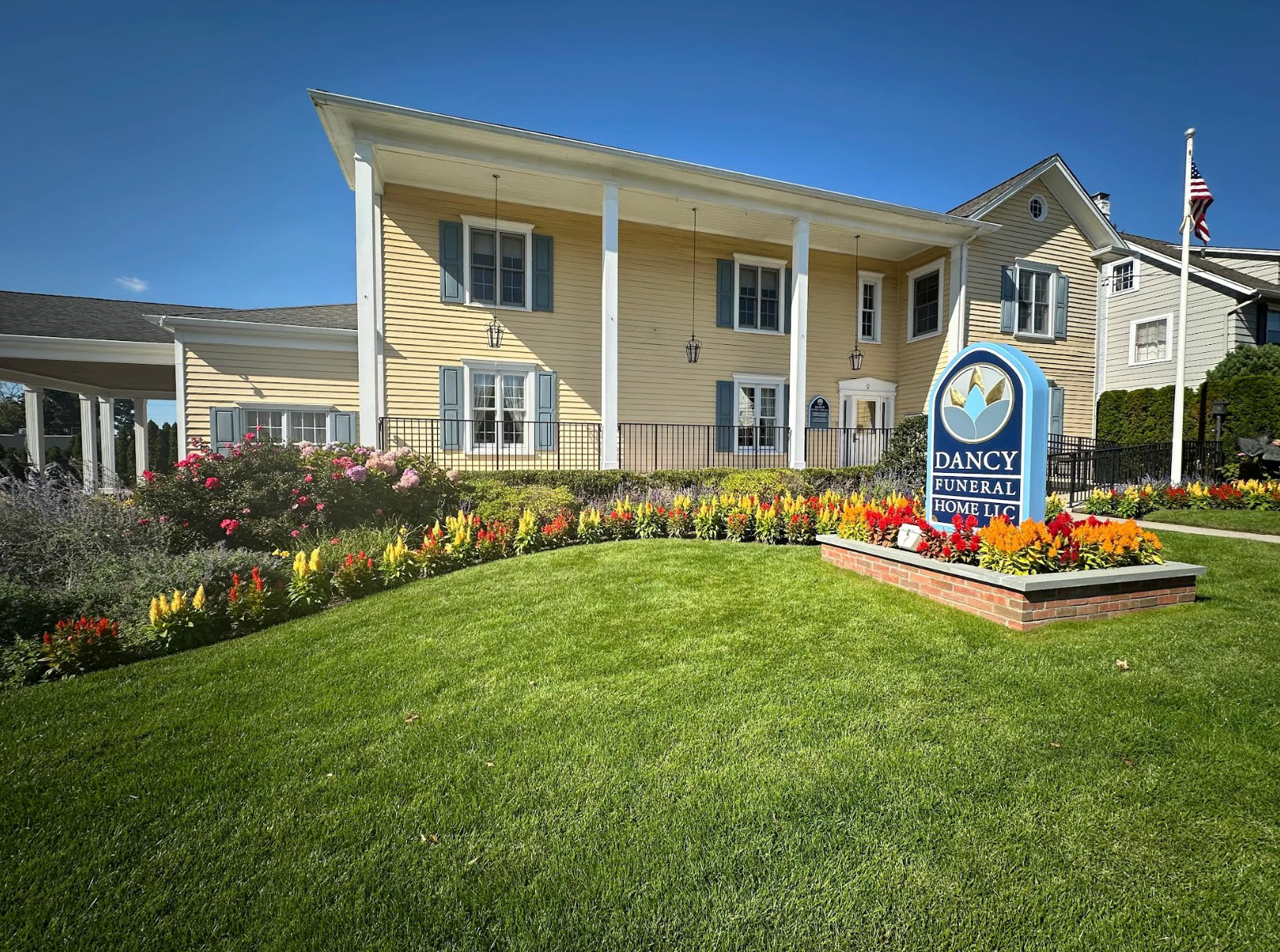 Yellow two-story building with porch, flower beds, and a blue sign in front on a sunny day