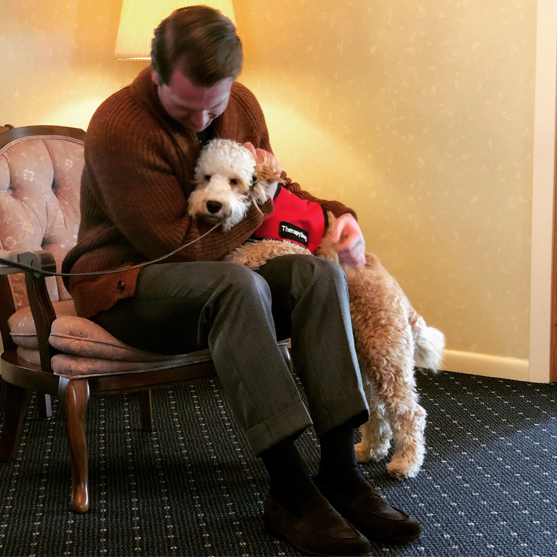 Man seated with a small white dog in a red vest, hugging it on a cushioned chair indoors.