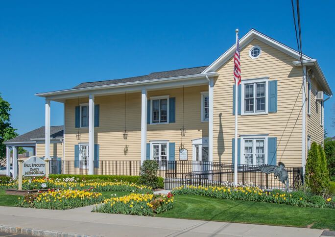 Yellow two-story house with white trim, blue shutters, and a flower garden on a sunny day