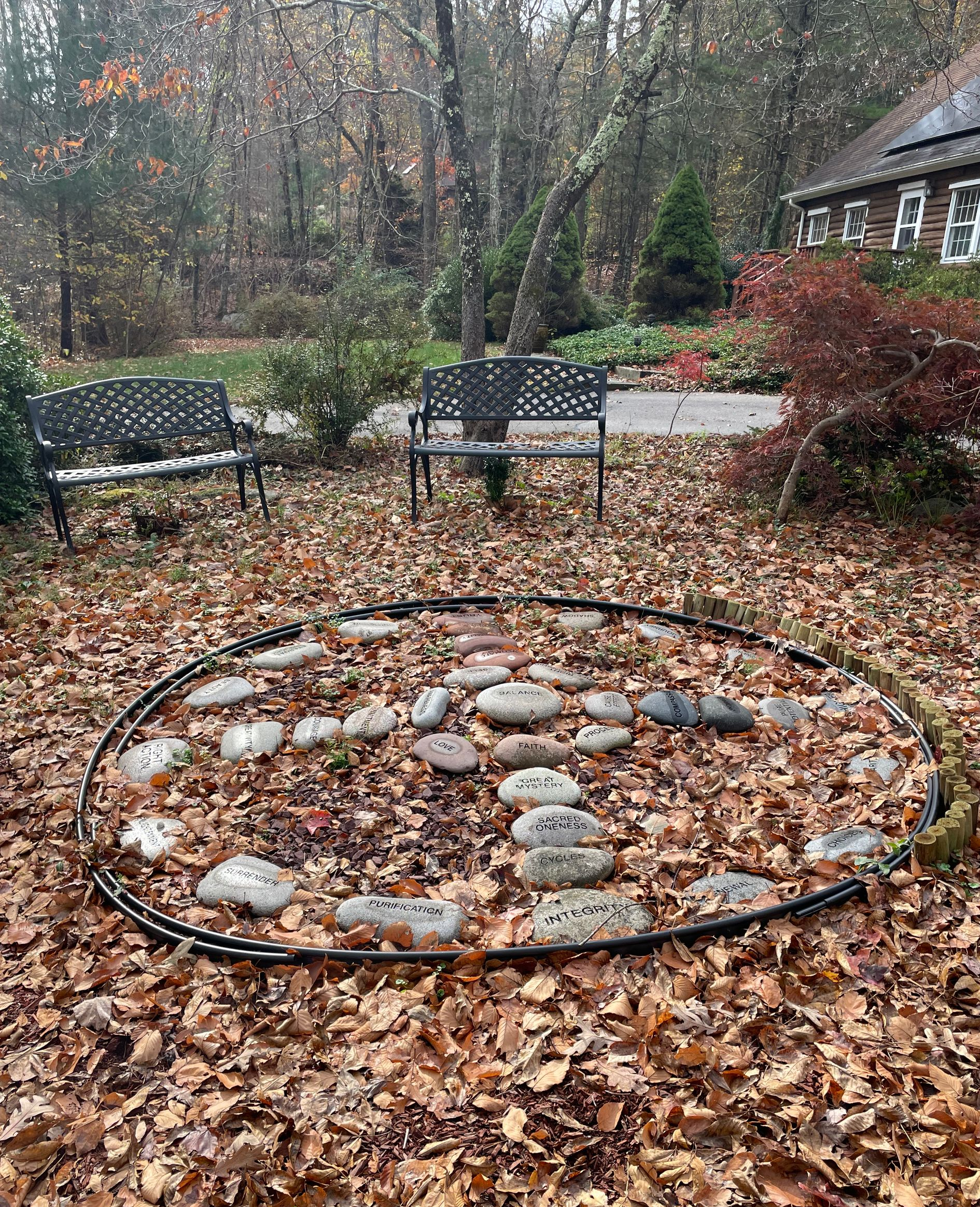 Circular stone fire pit in a leaf-covered backyard garden with benches and trees in autumn.