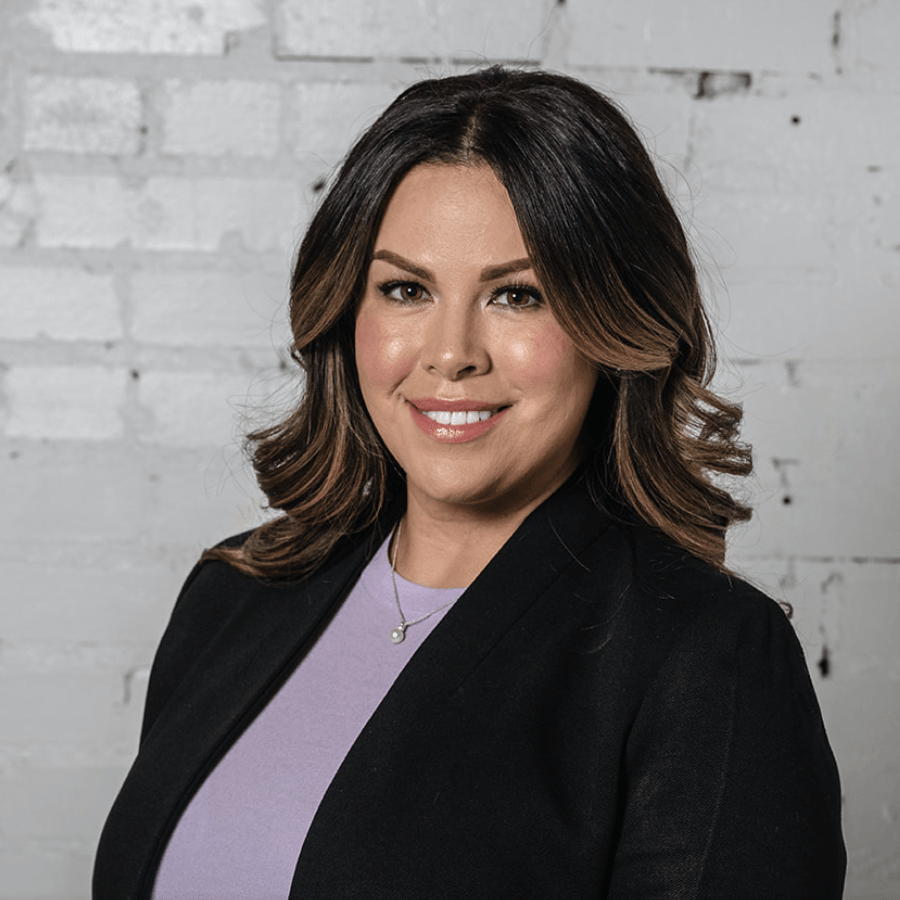 A woman in a black jacket and purple shirt is smiling in front of a white brick wall.