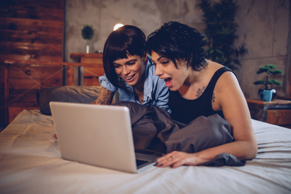 Two people smiling and leaning over a laptop on a bed in a cozy bedroom