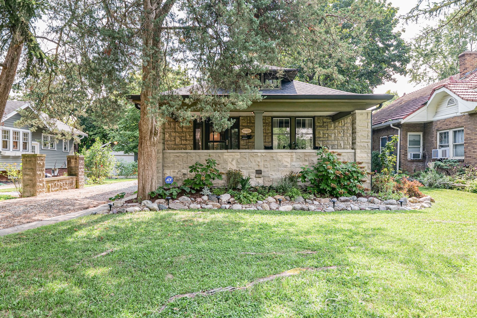 Single-story brick house with a front porch, large tree, and landscaped yard