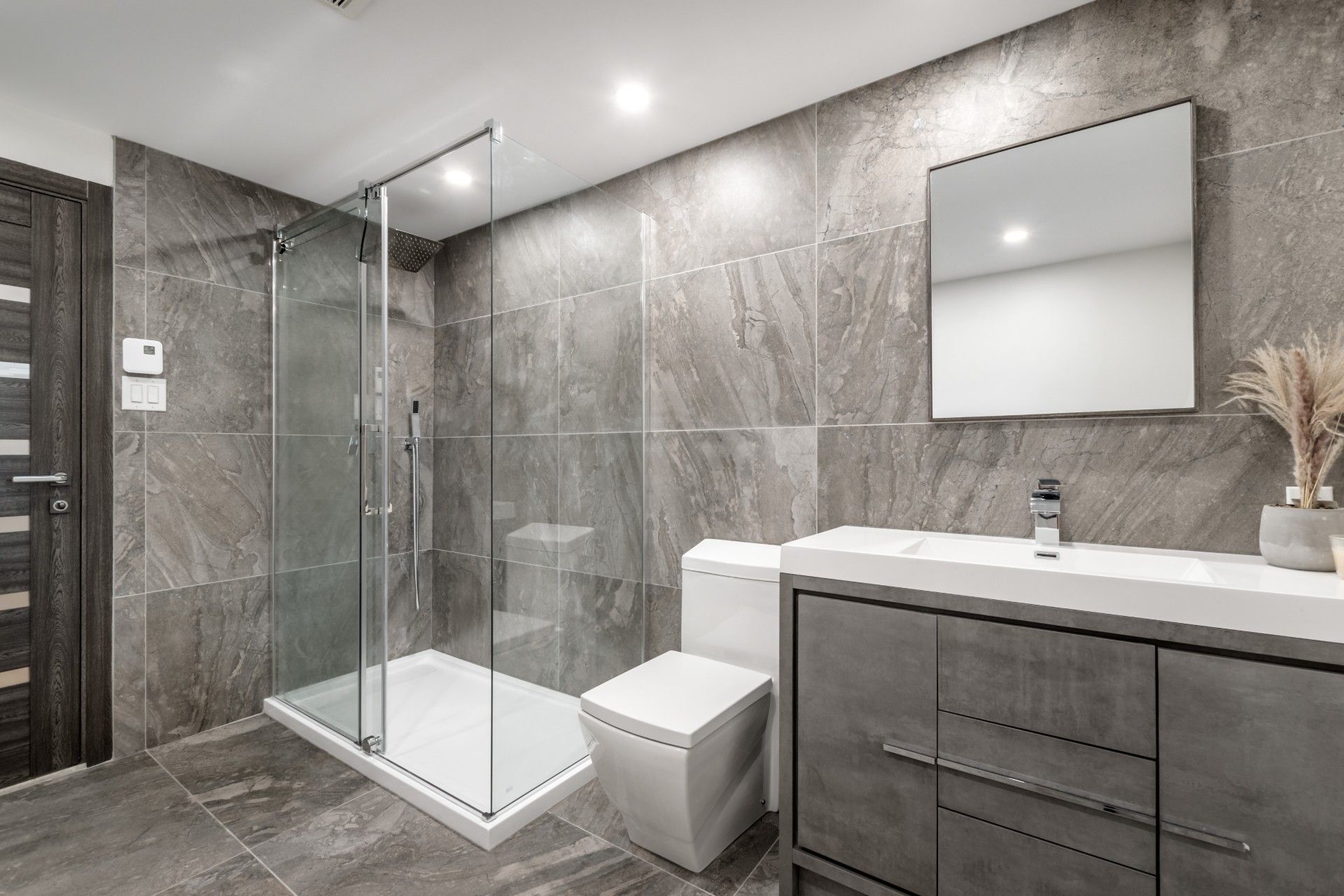 Modern gray bathroom with glass walk-in shower, toilet, vanity, and large mirror
