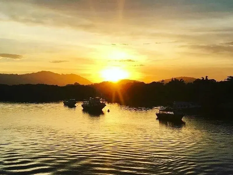 Pôr do sol dourado sobre um porto tranquilo, com silhuetas de vários barcos pequenos ancorados na água contra colinas distantes.