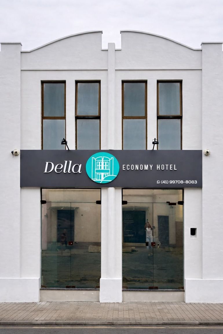Um edifício de hotel branco de dois andares chamado "Della Economy Hotel" com um logotipo circular verde-azulado e entrada de vidro no térreo.