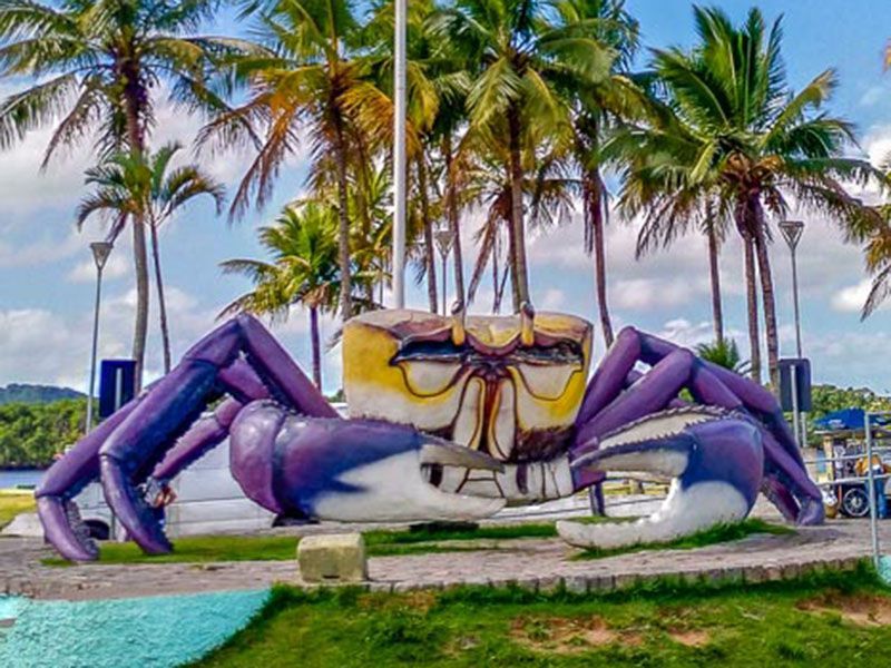 Uma grande escultura de caranguejo roxa e amarela se ergue em um parque tropical com palmeiras sob um céu azul brilhante.