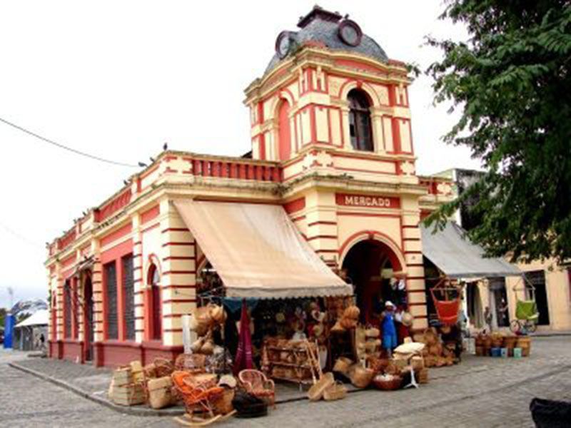 Um edifício histórico de mercado com uma torre de relógio laranja, onde se exibem tecidos e artesanato numa praça de calçada de pedra.