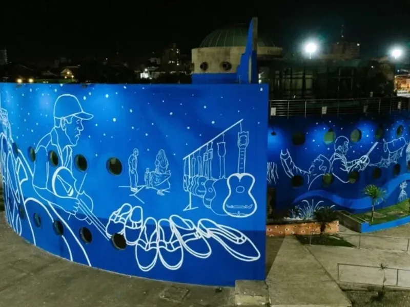À noite, uma parede curva exibe um mural azul com desenhos em linhas brancas de um músico, instrumentos e sapatos tradicionais.
