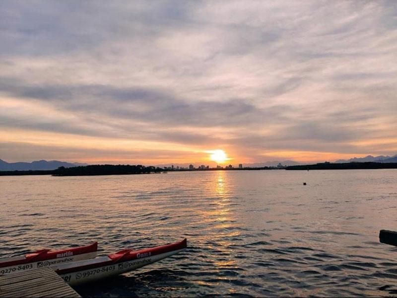 Um pôr do sol sobre uma superfície de água calma com uma canoa no cais em primeiro plano, emoldurado por colinas distantes e nuvens suaves.