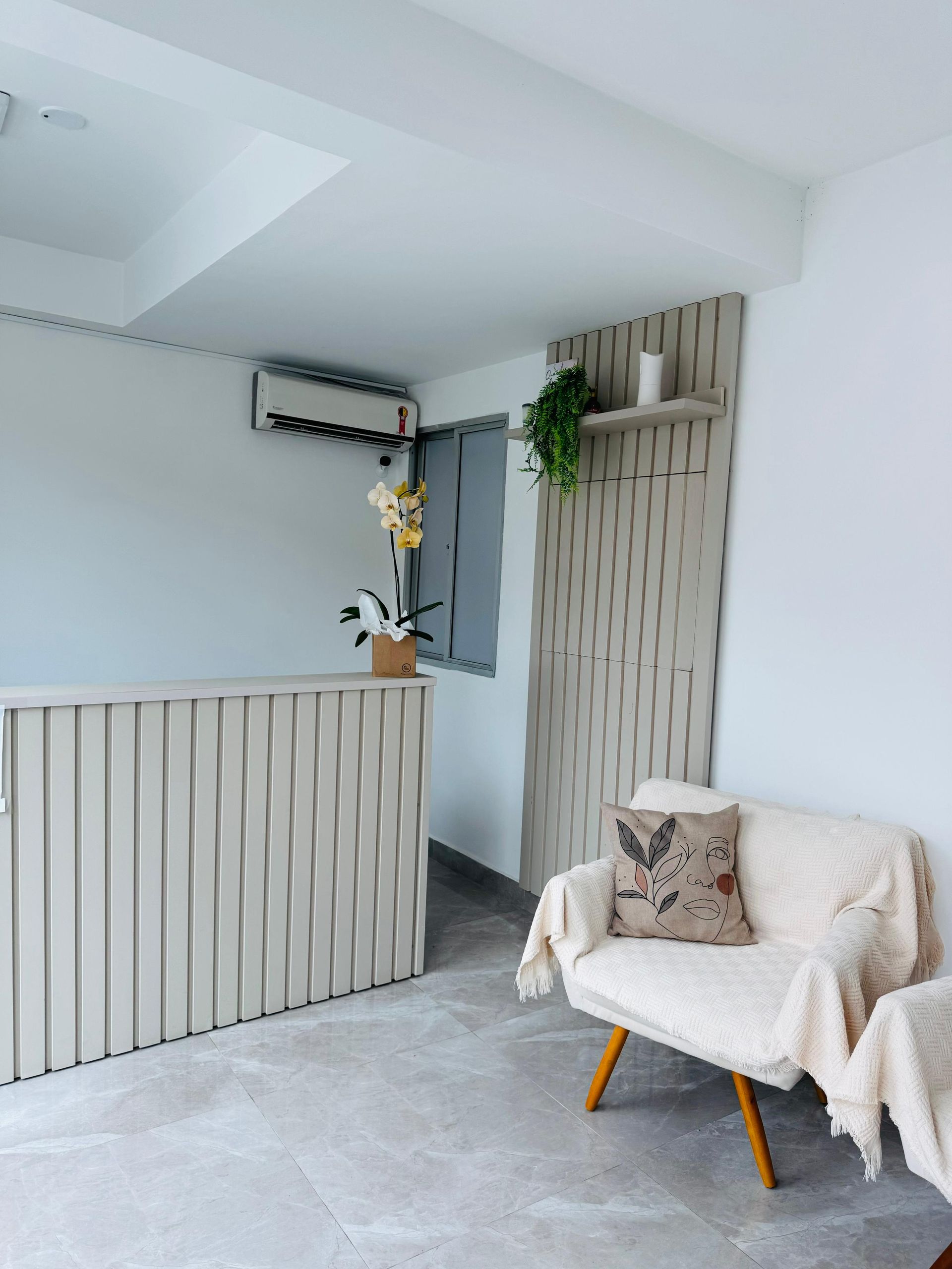Uma área de espera com um balcão de recepção em ripas de madeira clara, um painel decorativo na parede combinando com uma prateleira e uma poltrona cor creme.