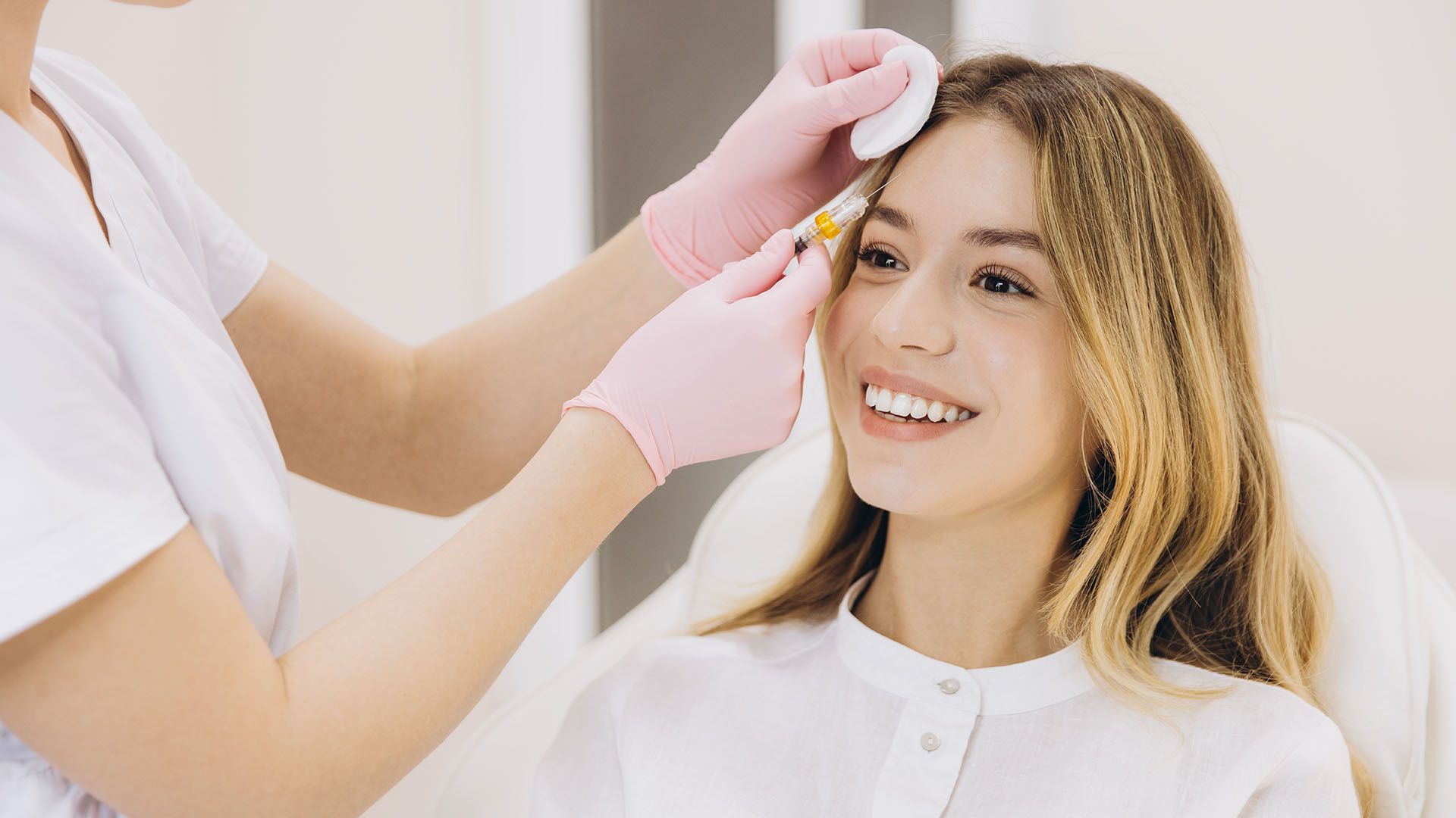 A smiling woman receives a facial injection in a medical setting; a gloved hand prepares the needle near her eyebrow.
