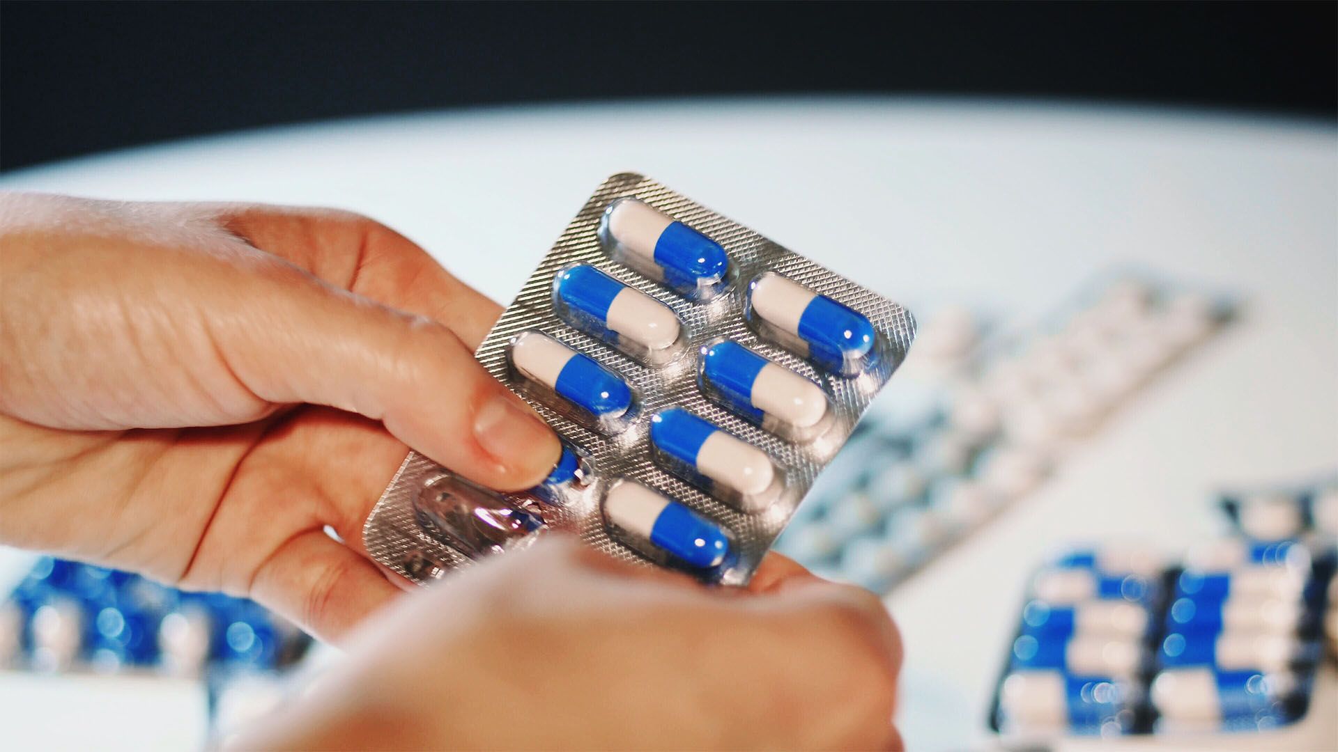 Hands holding a blister pack of blue and white capsules, with other packs in the background.