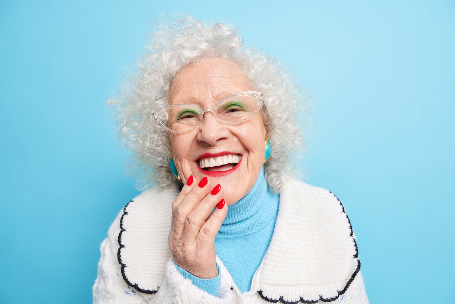 Smiling person with curly white hair, glasses, and red nails against a blue background