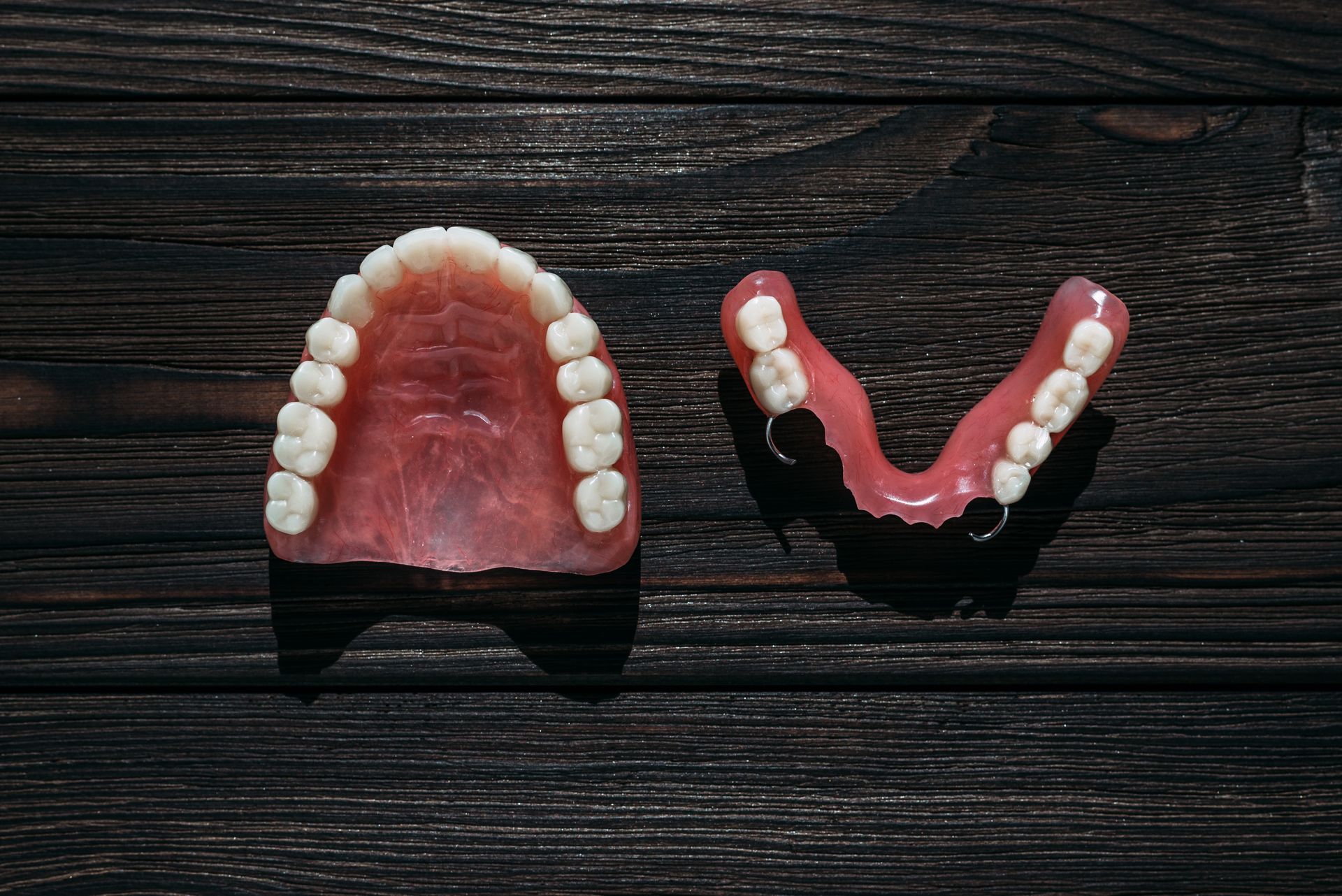 Two sets of dentures, one full upper denture and one lower partial, on a dark wooden surface
