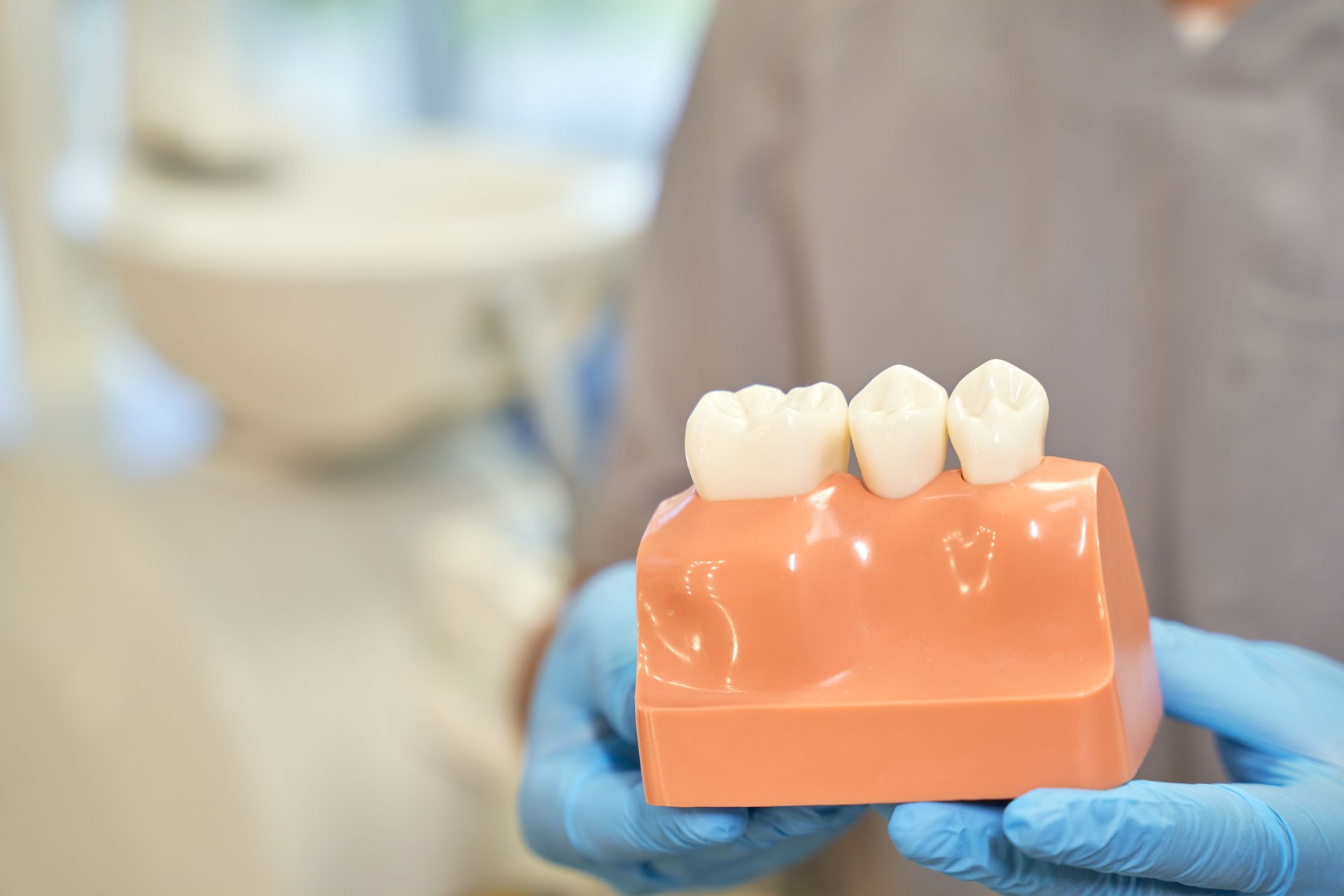 Dental model with three white teeth on a pink gum base, held by gloved hands in a clinic