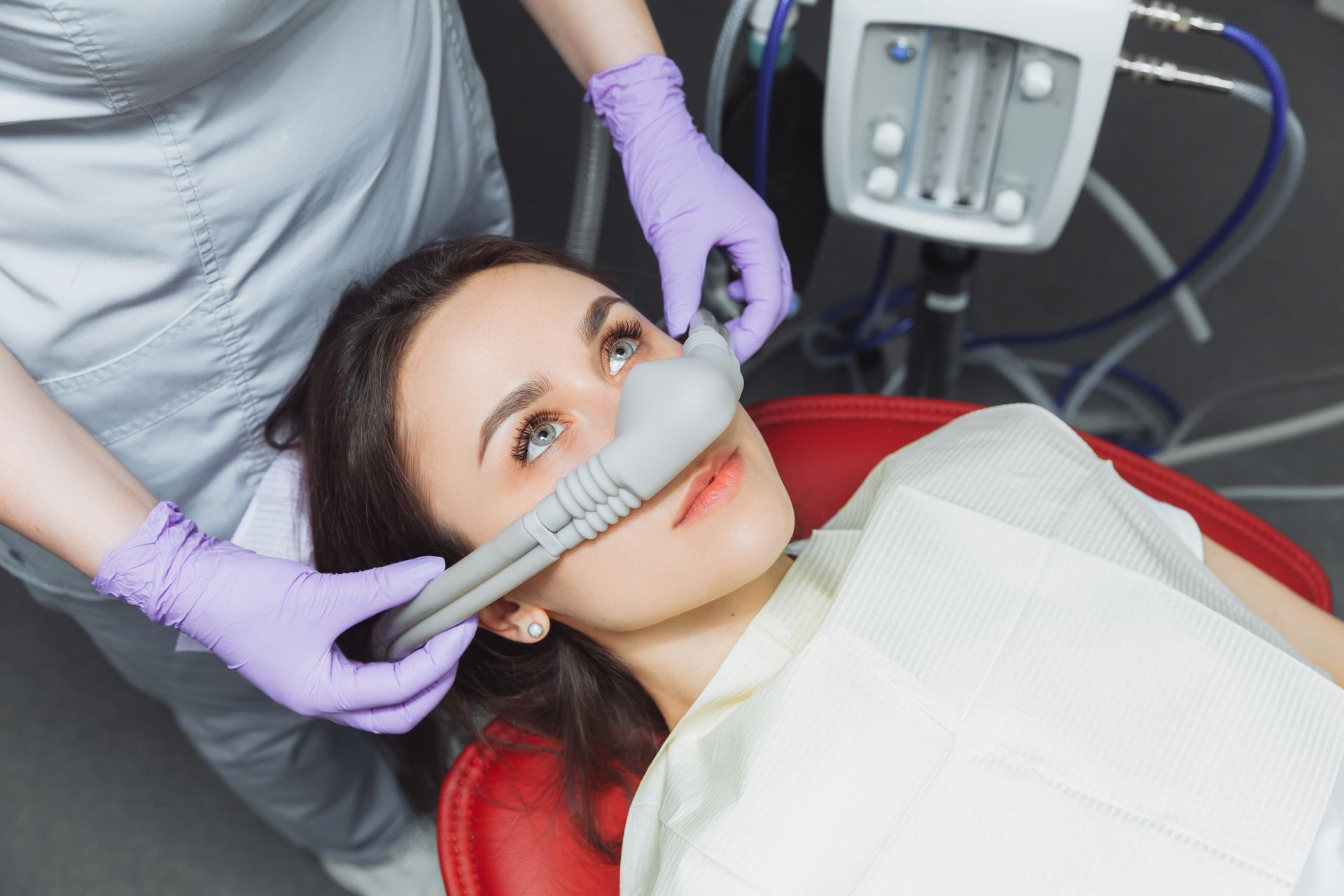 A dental professional in purple gloves places a nitrous oxide sedation mask over a patient’s nose in a clinical setting.