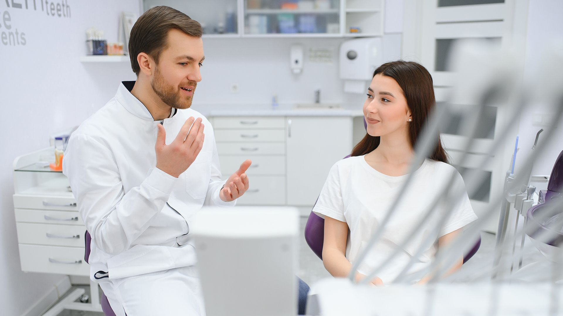A dentist in a white coat speaks to a patient in a dental chair in a bright office.
