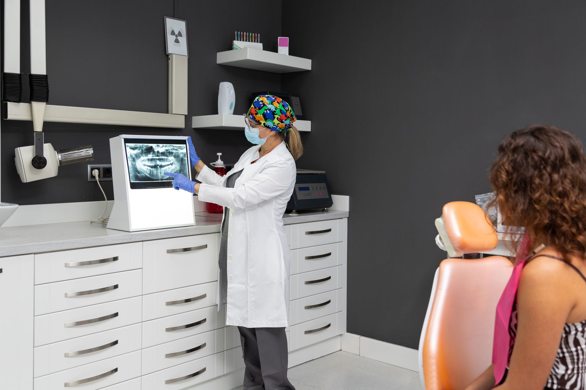 A dental professional in a white coat and mask explains a digital panoramic dental X-ray to a patient in a clinic.