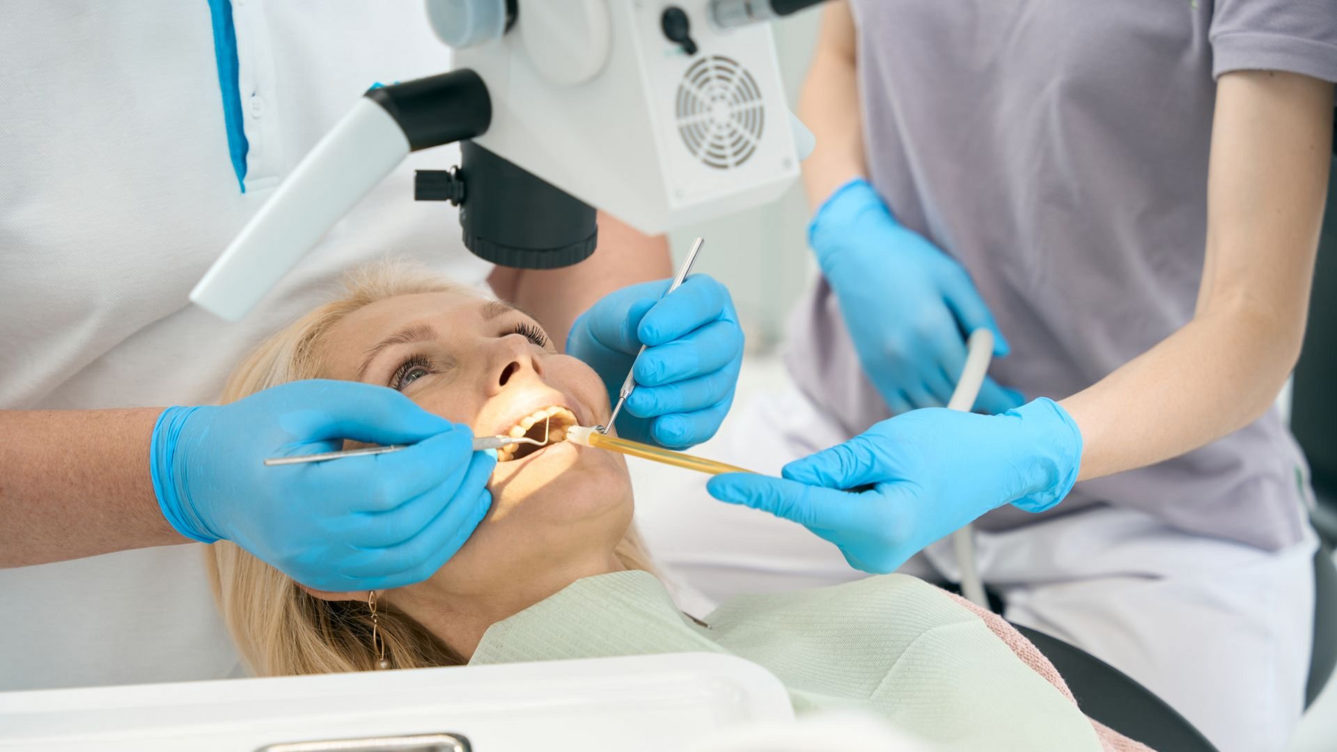 Dentist examining a patient's teeth with a dental microscope, assistant present.