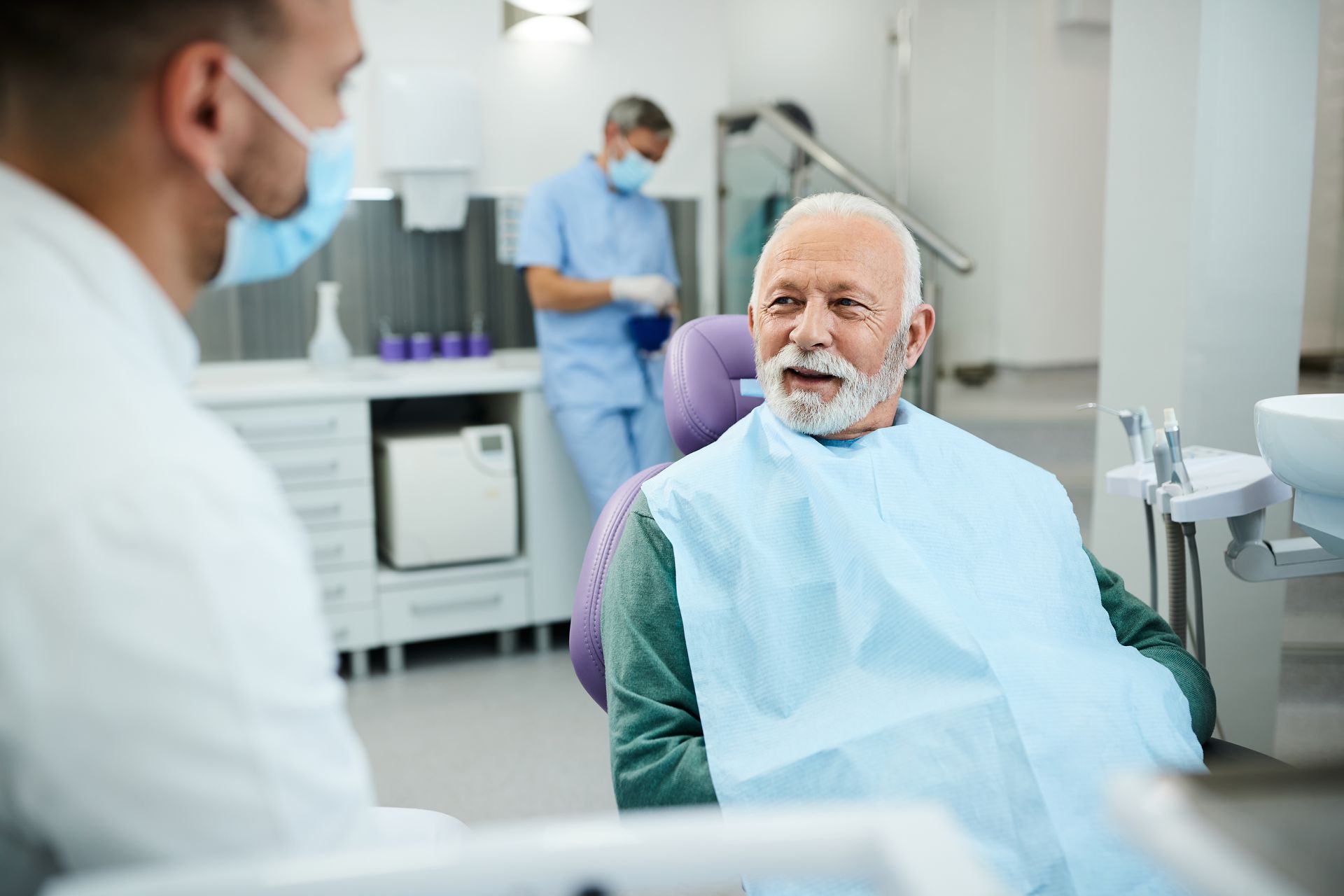 A dentist talks to a senior patient in a dental chair; a medical assistant in the background.