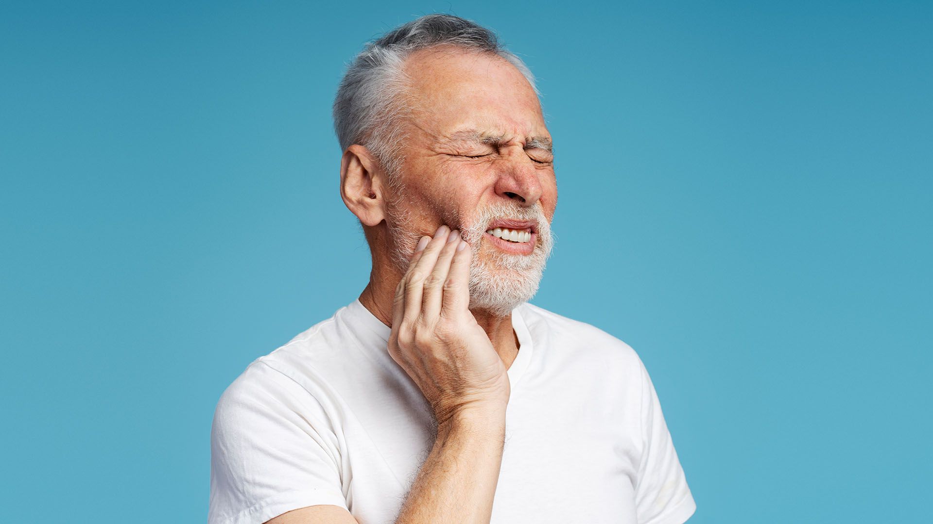 An older man with a grey beard, wearing a white shirt, clutches his jaw with a pained expression against a blue background.