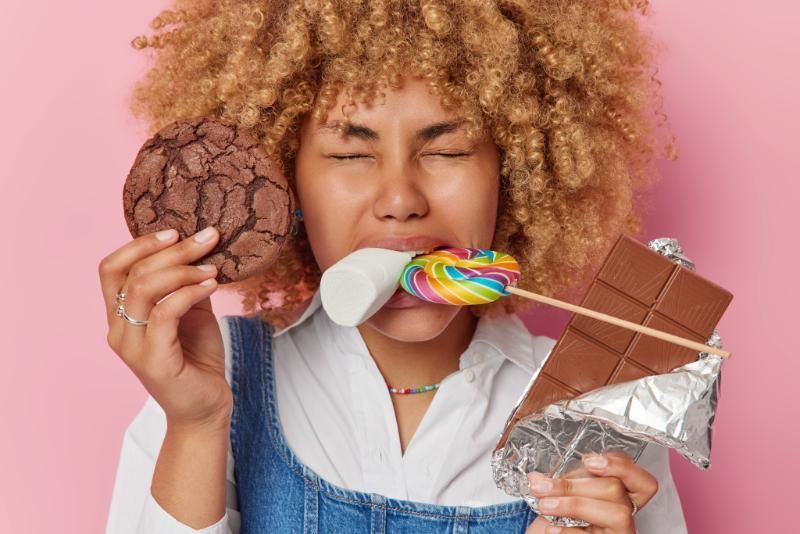 A woman is eating a cookie and a lollipop while holding a chocolate bar.