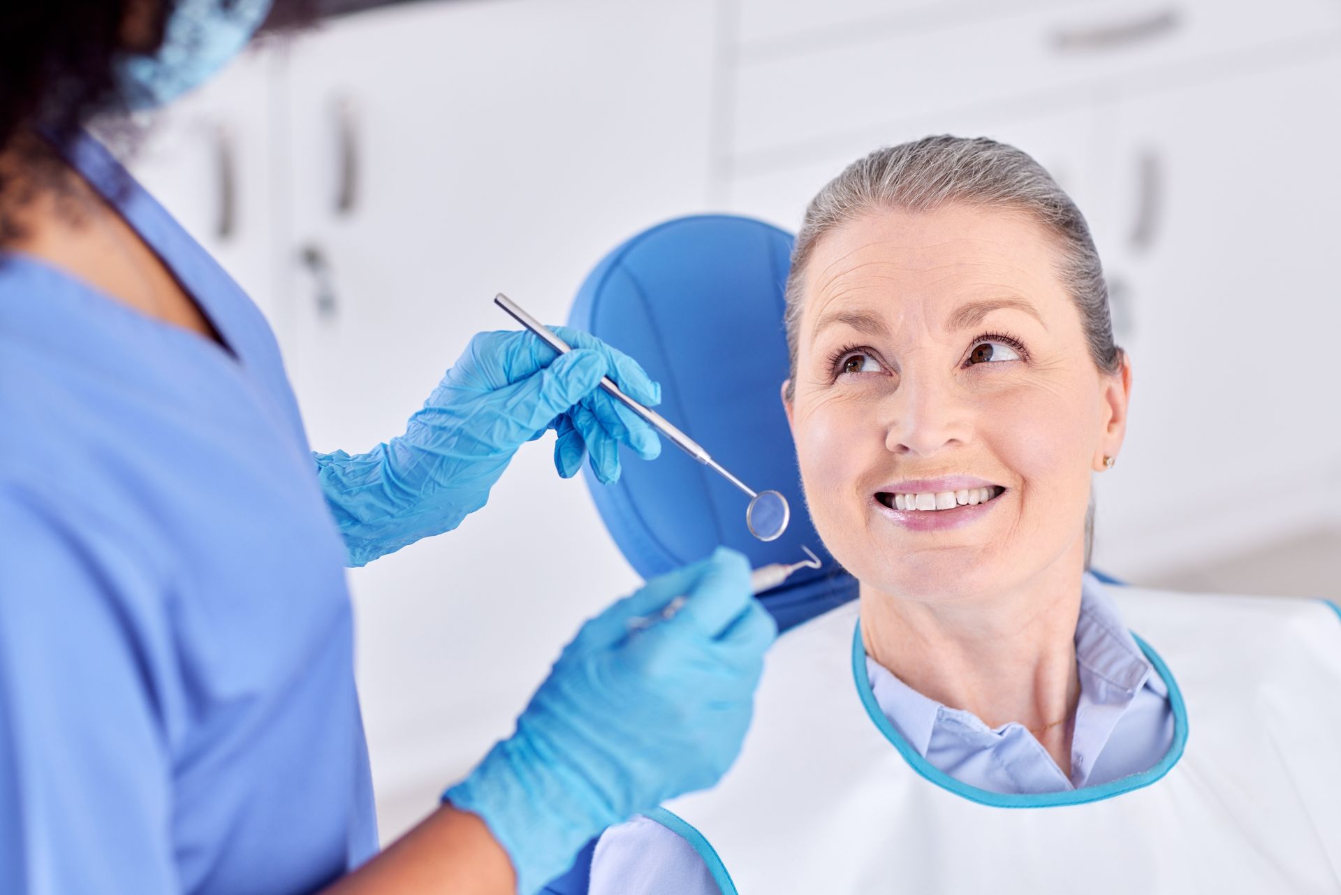 Woman in dental chair smiles as dentist examines teeth with instruments.