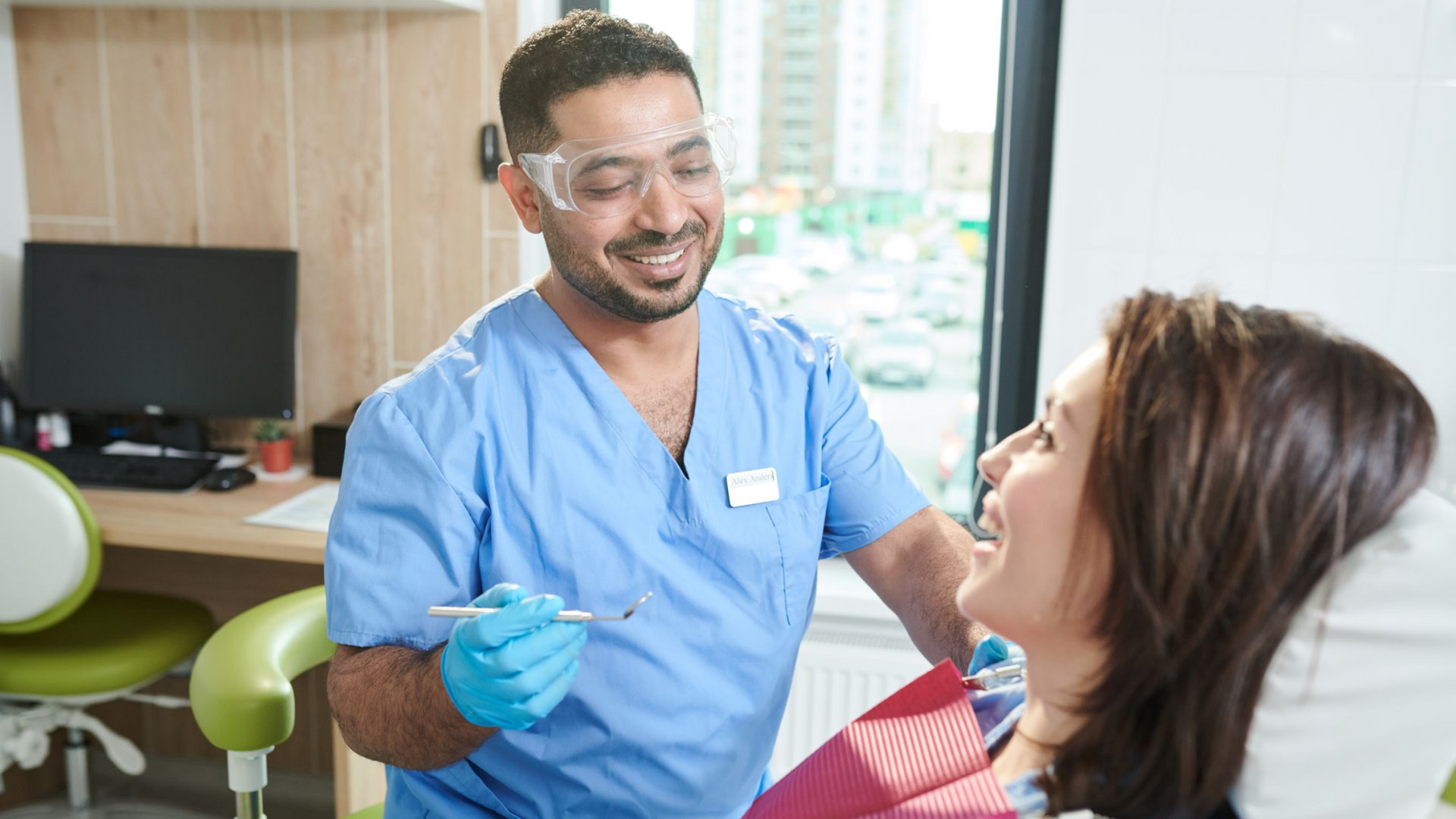 A dentist in blue scrubs examines a patient's teeth, smiling. The setting is a dental office.