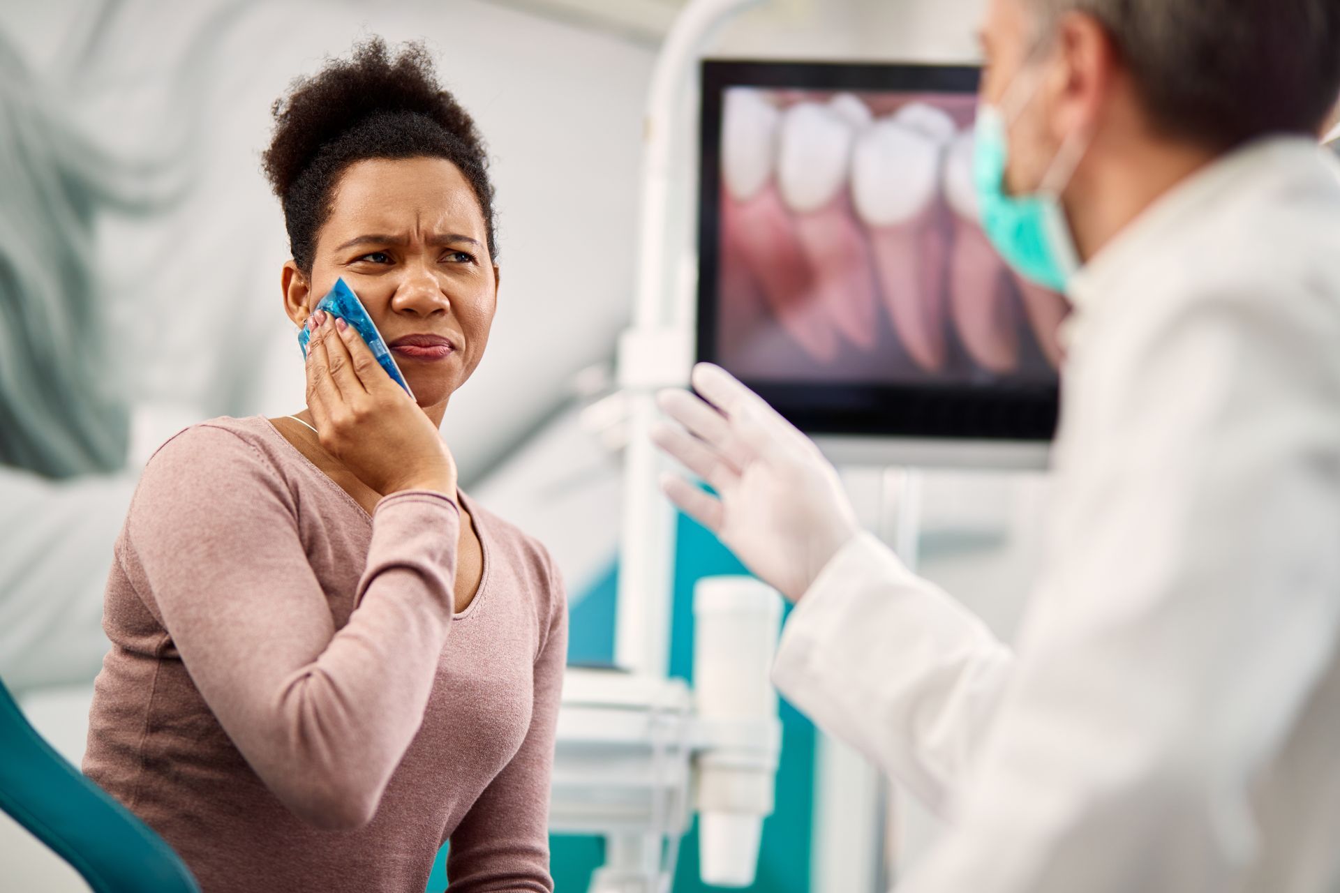 Woman in dental chair holding ice pack to cheek, looking pained. Dentist gesturing, dental screen visible.