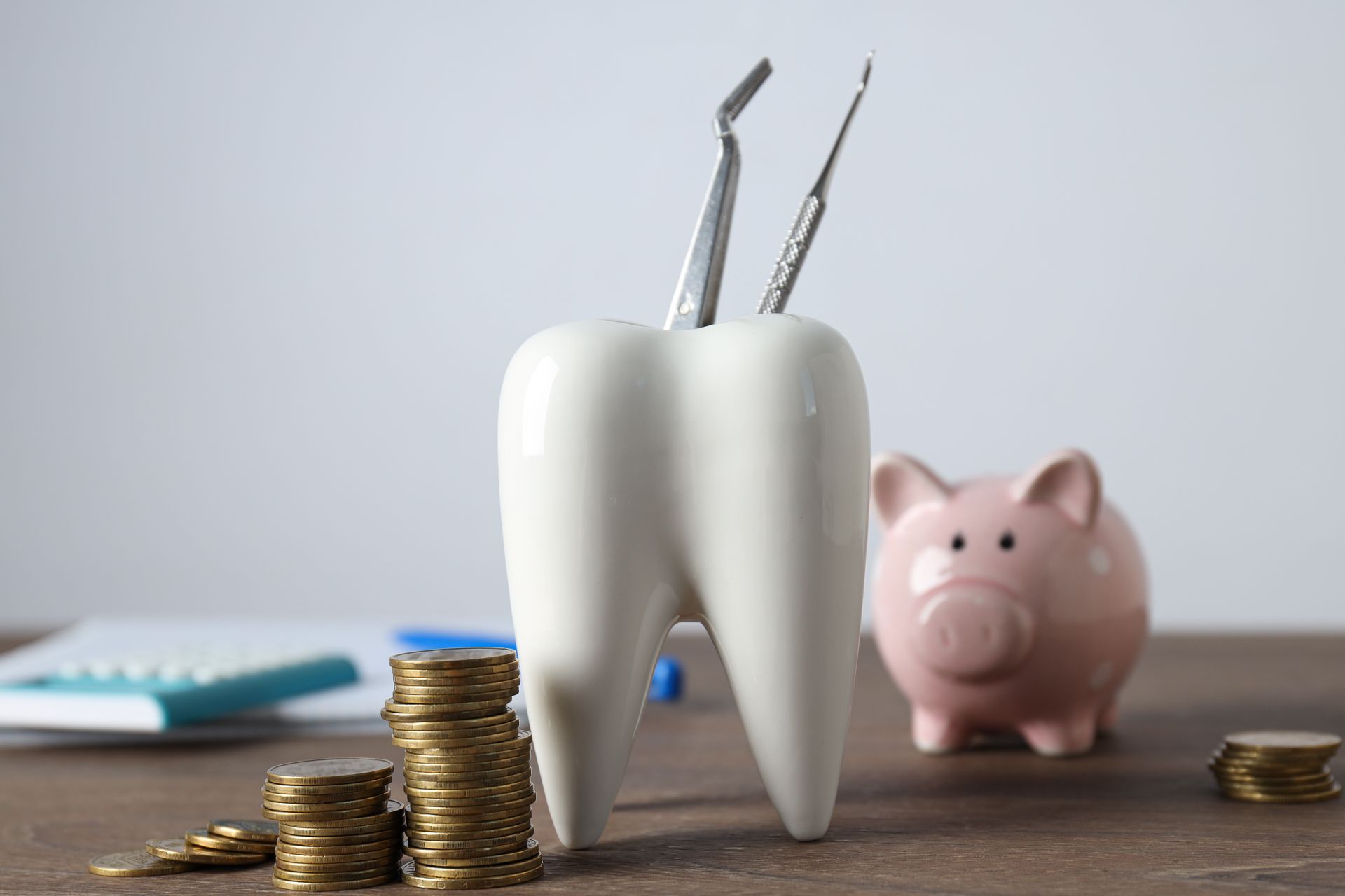 Tooth-shaped holder with dental tools, stacks of coins, and a piggy bank on a wooden surface.