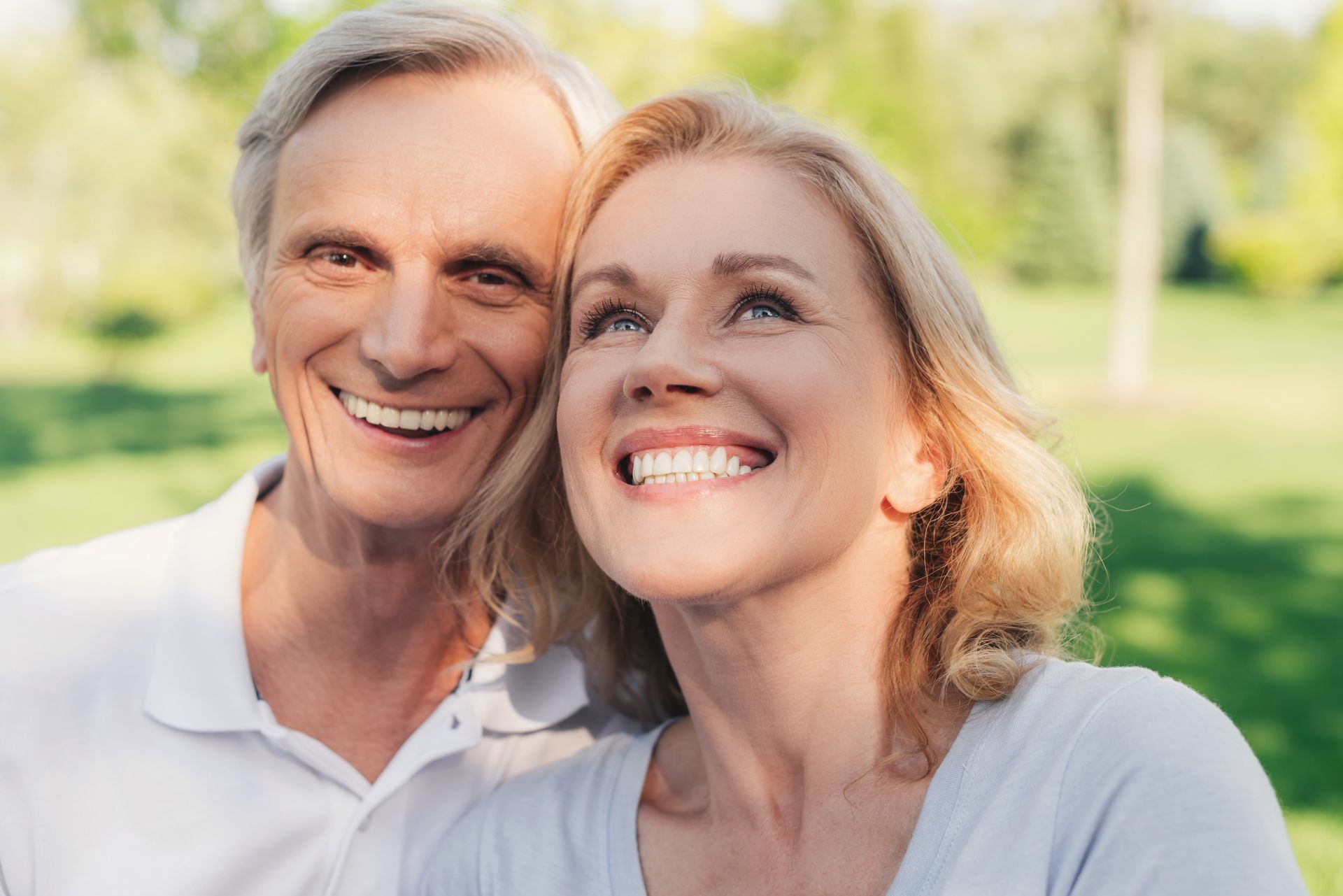 Smiling older couple outdoors, looking upwards with joy. Sunny day, green background.
