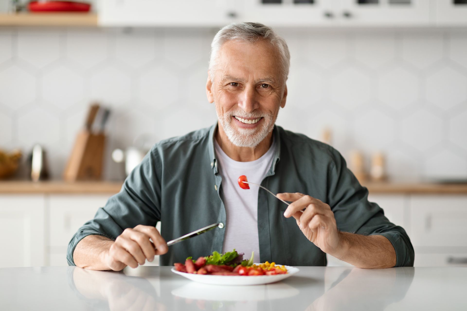 Smiling man at a table holding utensils, about to eat a salad in a bright kitchen