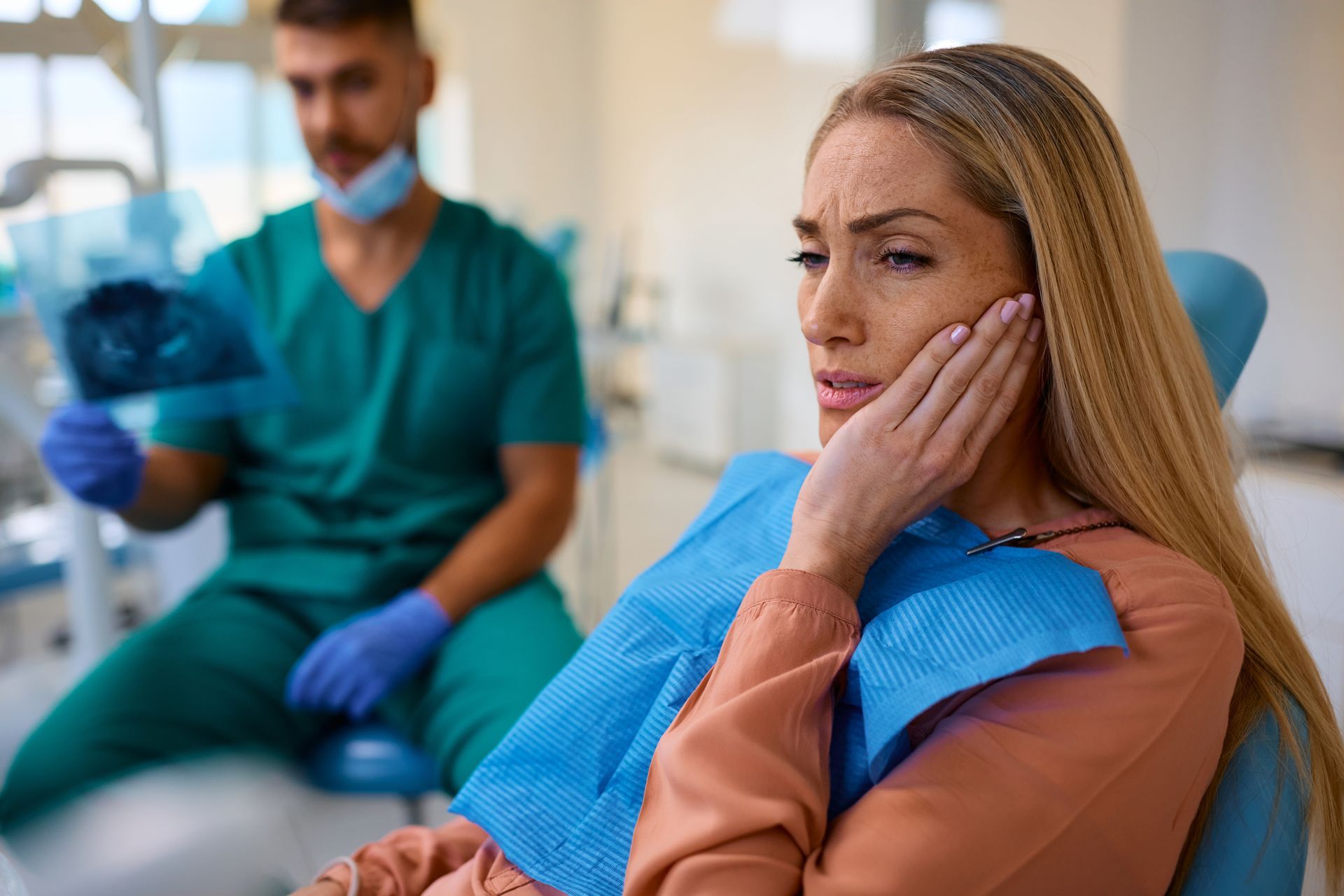 Woman sitting in a dental chair, holding her cheek while a dentist works in the background.