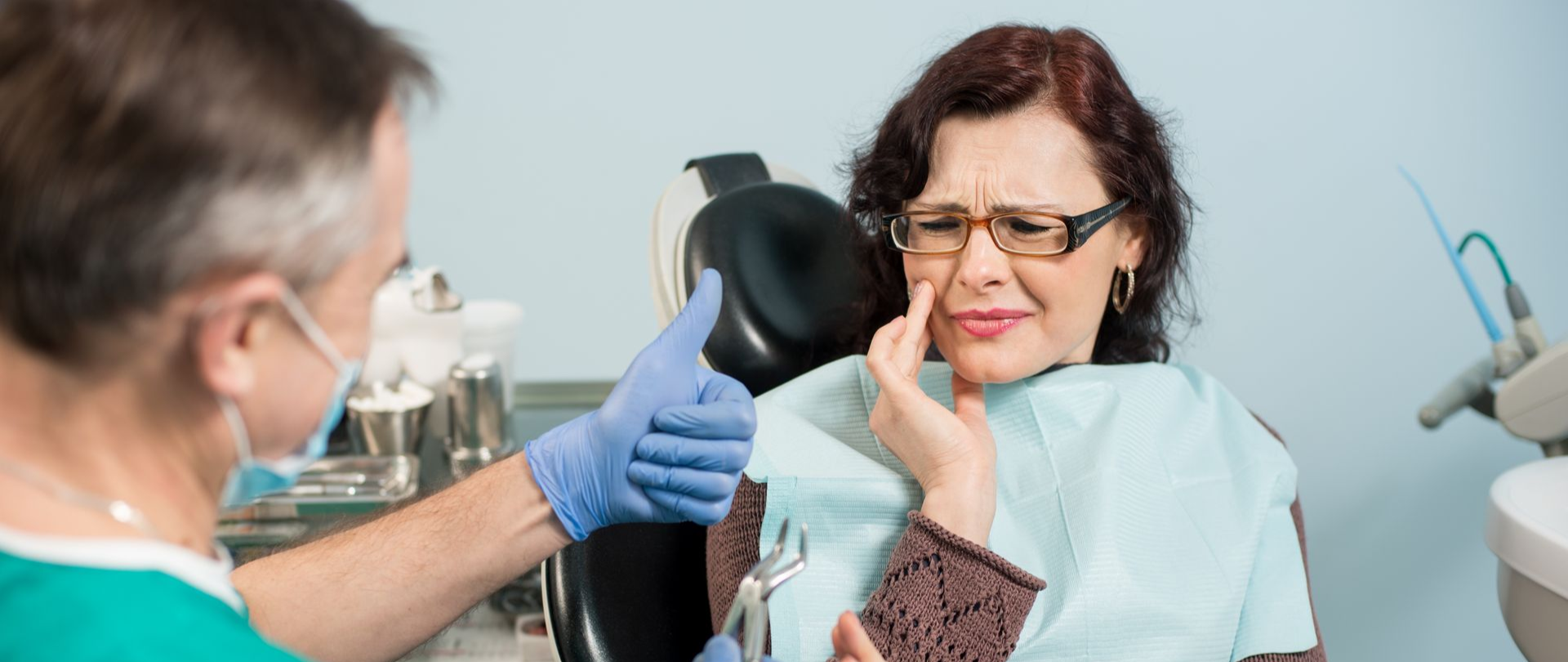A woman in a dentist chair is holding her cheek, showing pain, while a dentist preps a tool.